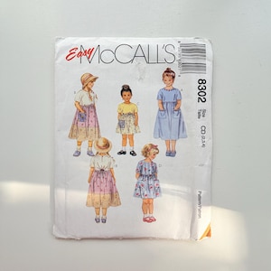 Può includere: Una busta per cartamodello Easy McCall's 8302 con illustrazioni di bambini che indossano vari stili di abiti. Il fronte mostra cinque diversi design di abiti con tasche, motivi floreali e colori a tinta unita, insieme alla gamma di taglie CD (2,3,4).