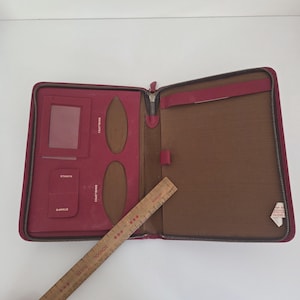 May include: A magenta zippered portfolio organiser with interior pockets labelled for envelopes and stamps, featuring a brown lining and a wooden ruler measuring approximately 30 cm in length placed diagonally across the open interior.