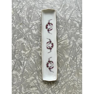 May include: A long, rectangular white ceramic serving dish with a gold trim. The dish is decorated with three floral wreath designs in shades of purple and green. The dish is ideal for serving appetizers or small portions.
