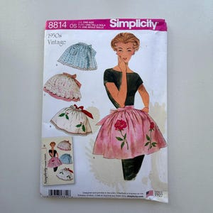 May include: A vintage Simplicity sewing pattern from the 1950s, featuring apron designs. The pattern shows illustrations of aprons with floral and patterned details. The pattern number is 8814, and the Simplicity logo is visible.