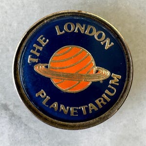 May include: A circular pin badge with a blue background and gold lettering that reads "THE LONDON PLANETARIUM." The badge features an orange planet with rings. The badge has a silver border.