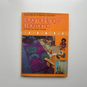 May include: A vintage book with an orange cover, titled "Rugmaking & Macramé: A Step-by-Step Guide" by Anna North & Noreen Gladwin. The cover shows a purple rug with green tree designs and examples of macrame.