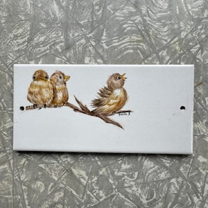 Può includere: Una piastrella rettangolare in ceramica bianca con un'illustrazione dipinta a mano di tre piccoli uccelli marroni appollaiati su un ramo. Due uccelli sono seduti insieme a sinistra, mentre un uccello con il becco aperto è a destra. Firmato Berta.