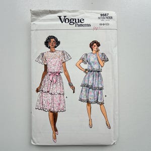 Könnte beinhalten: Vogue Patterns Schnittmusterumschlag mit Illustrationen von zwei Kleidern. Der Umschlag ist weiß mit dem Vogue-Logo und den Musterdetails. Ein Kleid ist rosa mit einer Schleife, das andere blau mit floralem Muster.
