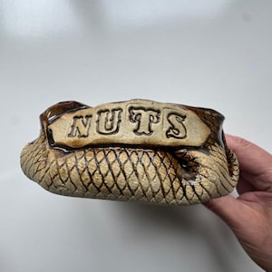 May include: A ceramic nut bowl with a textured, scale-like pattern in shades of brown and tan. The word "NUTS" is embossed across the top. The bowl is designed to hold snacks.