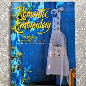 Peut inclure: Un livre intitulé "Romantic Embroidery" avec une couverture bleue et des lettres jaunes. La couverture présente une image d'un tablier blanc avec des broderies noires suspendu à une porte bleue. Le livre est un guide de projets de couture.