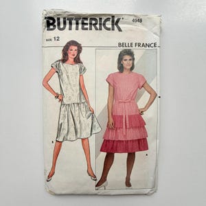 May include: A Butterick 4948 sewing pattern envelope for size 12 Belle France dresses. The front shows two illustrations: a white dress with a drop waist and a pink tiered dress with a tie waist, both featuring short sleeves and round necklines.