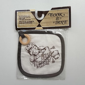 May include: A square off-white pot holder with a dark brown border and a wooden ring attached to the top corner. The centre features a black line drawing of a basket of bread with the text BREAD WORKS. It is in a clear plastic package with a header card labelled COOK & DINE.