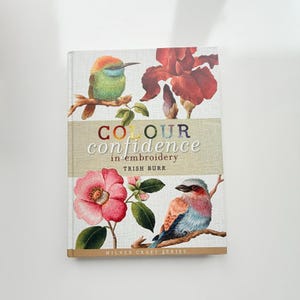 May include: A hardcover book titled Colour Confidence in Embroidery by Trish Burr. The cover features a green and orange bird on a branch, a large red flower, a pink camellia, and a blue and orange bird, all set against a light fabric background.