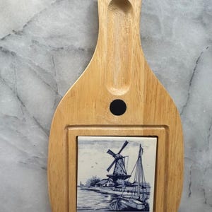 May include: Wooden cheese board with a handle and a decorative tile insert. The tile features a blue and white illustration of a windmill, boats, and a waterside village. The board is made of light-coloured wood.