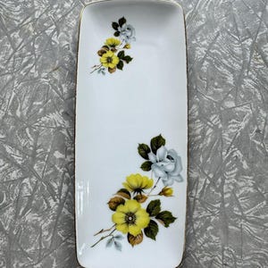 May include: A rectangular white ceramic serving dish with a gold rim. The dish is decorated with yellow and blue floral clusters and green leaves. The dish is approximately 8 inches long and 4 inches wide.