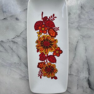 Puede incluir: Un plato rectangular de cerámica blanca con un diseño floral. El diseño presenta flores y hojas naranjas, amarillas y rojas. El plato es rectangular con esquinas redondeadas.