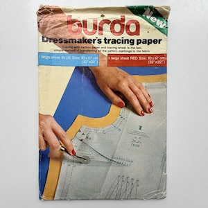 Op de afbeelding: Een Burda kopieerpapierverpakking voor kleermakers met een afbeelding van een hand die een kopieerwieltje op een naaipatroon gebruikt. De tekst bevat "Dressmaker's tracing paper" en vermeldt één blauw en één rood vel van 83x57 cm.