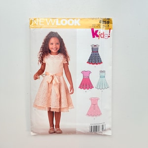 May include: A New Look sewing pattern envelope for kids, model 6359, featuring a photograph of a child wearing a peach-coloured lace dress with a satin ribbon sash. The envelope displays four illustrated dress variations in different colours and patterns.