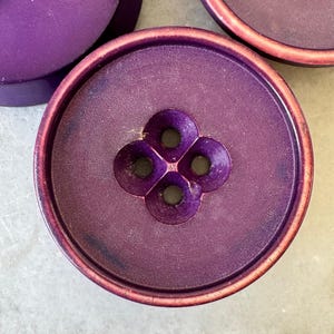 Peut inclure: Trois boutons ronds violets avec un bord extérieur surélevé et un centre en retrait. Chaque bouton présente quatre trous disposés en forme de trèfle au milieu. La surface des boutons a une finition mate avec de légères variations de couleur.