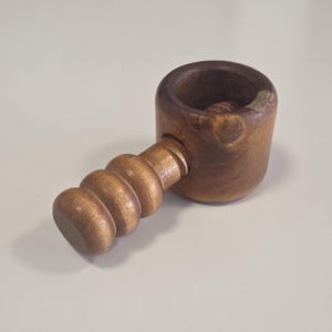 May include: A brown wooden nutcracker featuring a cylindrical base with a hollow centre and a threaded screw mechanism. The handle is shaped with three rounded, stacked segments, and the wood displays a natural grain pattern with a small chip.