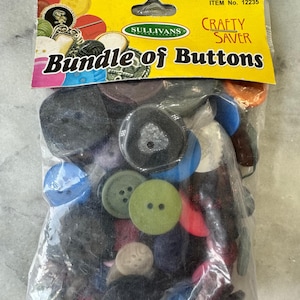 May include: A clear bag containing a selection of buttons in various colours and sizes. The yellow header reads "Bundle of Buttons" and "Crafty Saver". Buttons include blue, green, black, and red. The net weight is 115g.