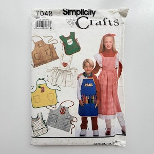 May include: A Simplicity Crafts sewing pattern envelope, number 7048, featuring illustrations of various aprons for children. The cover displays a child wearing a blue apron with the name Paul and a crayon graphic, alongside a red and white gingham apron.