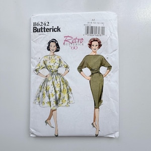 May include: A Butterick B6242 sewing pattern envelope featuring two dress designs. View A shows a floral print dress with a full skirt, while View B displays a solid olive green dress with a fitted skirt. Both designs feature boat necklines.
