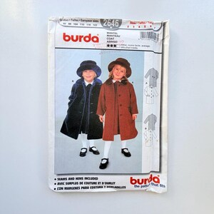 Könnte beinhalten: Ein Burda 2646 Schnittmusterumschlag für einen Mantel. Die Vorderseite zeigt Illustrationen von zwei Kindern in Mänteln und Hüten, eines in Dunkelblau und eines in Rostbraun. Der Text enthält Burda, Mantel, Manteau, Coat, Abrigo und Größenangaben.