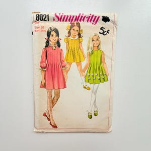 Op de afbeelding: Een vintage Simplicity naaipatroon-envelop, nummer 8021, voor een meisjesjurk in maat 10 met een borstomvang van 72,4 cm. De hoes bevat illustraties van drie jurken in roze, geel en groen, met een prijs van 5 cent gemarkeerd in zwarte inkt.