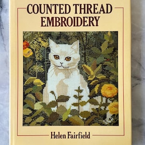 May include: A book titled "COUNTED THREAD EMBROIDERY" by Helen Fairfield. The cover features a cross-stitch image of a white cat amidst green foliage and yellow flowers. The book has a cream-colored background with red borders.