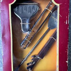 May include: Deluxe Bar Set in a red box. The set includes a bottle opener, tongs, a spatula, and a bar knife. The tools have silver metal parts and dark wood or black handles. The box has a gold and yellow interior.