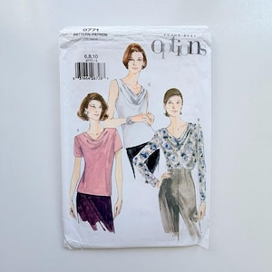 May include: A Vogue Easy Options sewing pattern envelope, number 9771, featuring illustrations of three tops with cowl necklines. The designs include a short-sleeved pink top, a sleeveless white top, and a long-sleeved blue floral print top.