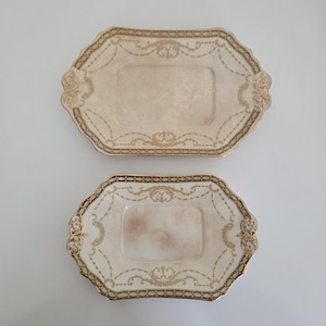 May include: Two rectangular ceramic trays with scalloped edges, featuring a cream-coloured centre and a decorative gold border with intricate filigree patterns and draped garland motifs. The trays are arranged vertically, one larger than the other.