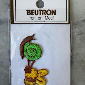 May include: A Beutron Iron on Motif patch featuring an embroidered snail with a brown body and a bright green spiral shell, positioned above a yellow and brown bumblebee. The patch is sealed in clear plastic packaging with a white header card.