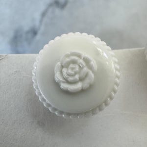 May include: A round, white milk glass knob featuring a raised, sculpted rose design at the centre. The edge of the knob is detailed with a decorative, beaded rope-like texture that encircles the entire circumference of the circular piece.