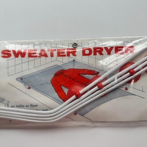 May include: A white plastic sweater dryer frame with red accents, packaged in a clear plastic bag. The packaging features the text "SWEATER DRYER" in red capital letters and an illustration of a red sweater resting on a mesh drying surface.