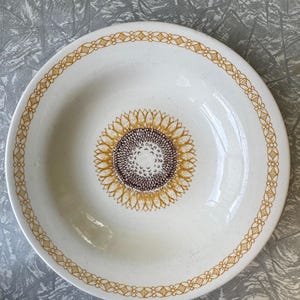 Peut inclure: Bol en céramique blanche avec un motif de tournesol au centre. Le tournesol a un centre marron et des pétales jaunes. Le bord du bol présente un motif floral jaune répété. Le bol est probablement destiné à servir des aliments.