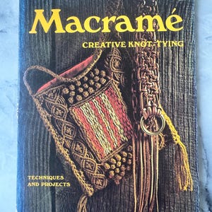 Könnte beinhalten: Ein Vintage-Buchcover mit dem Titel "Macramé" und dem Untertitel "Creative Knot-Tying". Das Cover zeigt eine Makramee-Tasche mit rot-weißen Streifen und einen Makramee-Riemen. Der Hintergrund ist eine Holzmaserung.