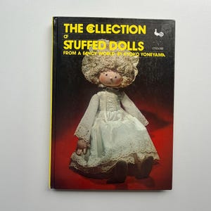 May include: A vintage book titled "The Collection of Stuffed Dolls" by Ryoko Yoneyama. The cover features a doll in a white dress with lace details and a bonnet. The book has a black cover with yellow edges.
