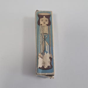 Peut inclure: Une boîte en carton rectangulaire avec un fond bleu et une illustration graphique d'un personnage stylisé aux cheveux brun foncé. La tête du personnage forme le haut d'un sablier, avec un minuteur en verre transparent visible à l'intérieur.