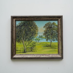 May include: A framed landscape painting featuring two trees on a green field, with a body of water in the background. The sky is light blue. The frame is silver and brown.