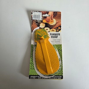 May include: A yellow plastic pineapple peeler with a textured pineapple-shaped body and a central handle, mounted on a cardboard backing card. The packaging features the text Pineapple Peeler, Reko Pty Ltd, and instructions for use.