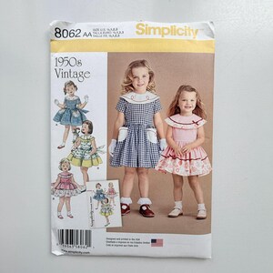 May include: A Simplicity 8062 sewing pattern envelope for 1950s vintage style dresses. The front features illustrations and photographs of children wearing gingham and floral dresses with large collars and pockets, sized US 1/2, 1, 2, 3.