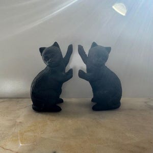 May include: Two black cat ornaments face each other, with their paws raised. The cat figurines are in a seated position. They are made of a dark material and are set against a light background.