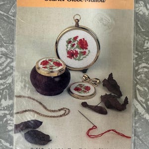 Op de afbeelding: Een knutselpakket getiteld "Scarlet Globe-Mallow" met kruissteekontwerpen. De afbeelding toont een kruissteek fob broche, een kleine houten kom met een kruissteekdeksel en een miniatuur messing frame. De set bevat een naald en rode draad.