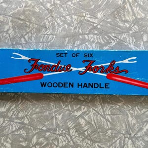 May include: A blue box displaying "Set of Six Fondue Forks Wooden Handle" in red and white. The box shows two fondue forks with red handles. The background is a textured, light grey surface.