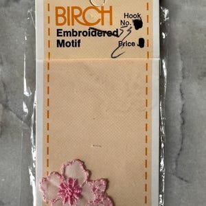 May include: A Birch brand embroidered motif in the shape of a light pink flower with a darker pink center, attached to a rectangular beige cardboard backing card with orange dashed borders, enclosed in a clear plastic protective sleeve.