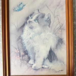 May include: A framed print of a fluffy white and gray cat with blue eyes looking up at a blue butterfly. The artwork has a soft, pastel color palette and is signed "KATHY" in the lower right corner. The frame is a warm, medium brown.