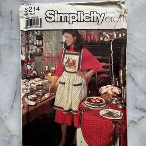 Op de afbeelding: Een vintage Simplicity Crafts patroon. De omslag toont een vrouw die een kerstthema schort draagt. De schort is wit met een rood en groen kerstontwerp en zwarte stippen. Het patroon bevat ook tafelaccessoires.