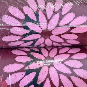 May include: Two rolls of burgundy fabric are wrapped in clear plastic. Each roll features a large, stylized flower design with pink petals and black accents. The floral pattern is repeated across the surface, creating a decorative effect.