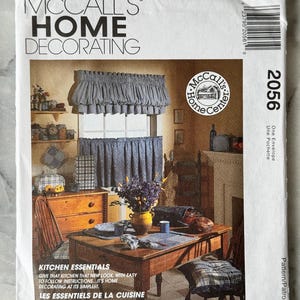 Op de afbeelding: Een McCall's Home Decorating patroon envelop met een keukenscène met gordijnen, een tafelsetting en tekst. De tekst bevat "Kitchen Essentials" en "Les Essentiels de la Cuisine". De envelop heeft een vintage esthetiek.