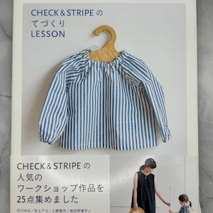 以下が含まれることがあります： 木製のハンガーにかけられた、日本語のテキストと青と白のストライプのトップスが表紙の書籍。書籍のタイトルは「CHECK & STRIPE のてづくり LESSON」です。表紙には、服や人々の画像も掲載されています。