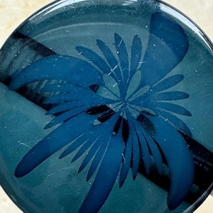 May include: A round, teal-colored glass object with a dark blue floral design. The design features a central cluster of petals surrounded by elongated, pointed leaves. The glass has a glossy finish.