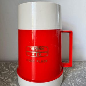 May include: A vintage red and white Thermos Brand food flask. The flask has a white top and base, with a red body and handle. The Thermos logo and "FOOD FLASK" are printed in gold. The flask is designed to keep food hot or cold.